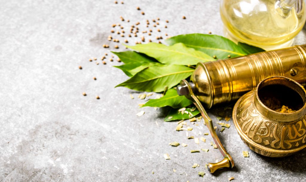 Antike Ayurveda-Mühle mit duftenden Gewürzen und Lorbeerblättern auf steinernem Tisch