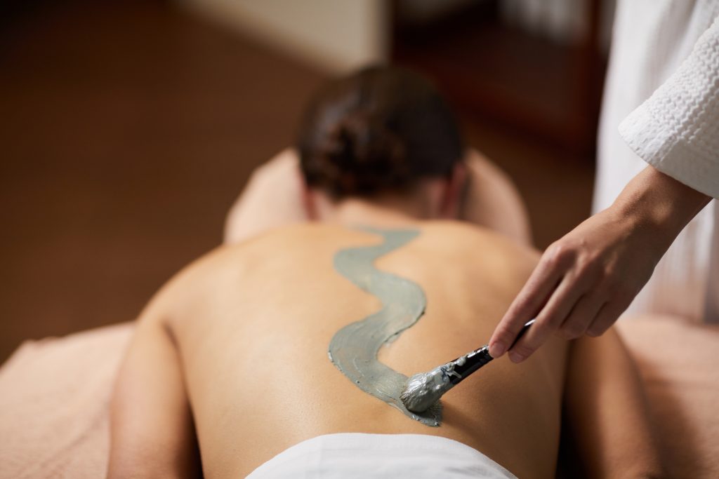 Kosmetikerin trägt Moor-Packung in Kurvenform auf den Rücken der Kundin während einer Spa-Behandlung auf